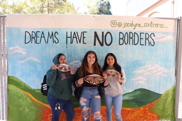 Three students holidng paper butterflies in front of the DREAMS HAVE NO BORDERS mural