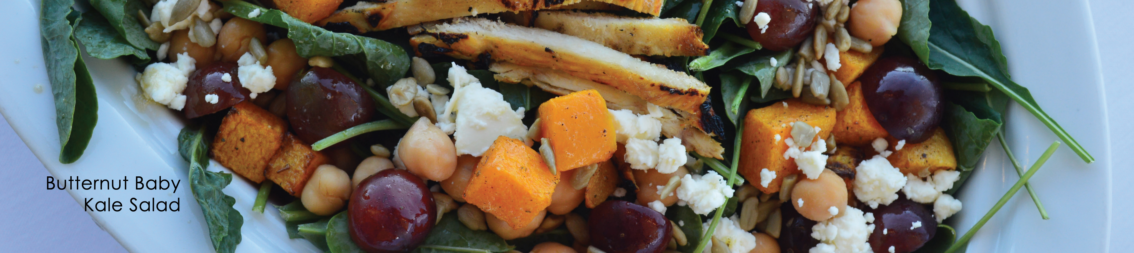 Butternut Baby Kale Salad. Nice salad plate with greens, croutons, grapes, cheese, and chicken on white plate.